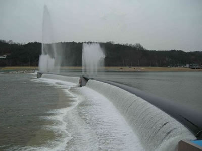 A curved black bookend rubber dam has overflow and there are many trees far away.